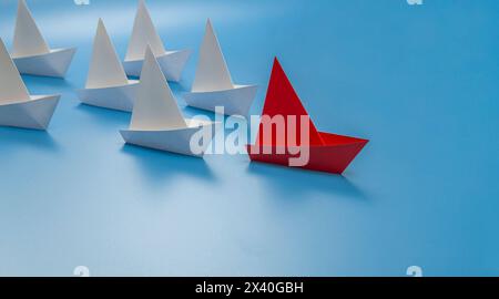 Rotes Papierboot schwimmt am Kopf einer Gruppe von weißen Papierbooten auf blauem Hintergrund. Führungskonzept. Stockfoto