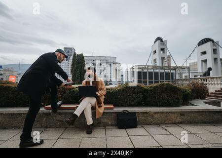 Zwei Geschäftsleute diskutieren im Freien mit einem Laptop. Moderne Stadtumgebung bildet den Rahmen für diese geschäftliche Interaktion. Stockfoto