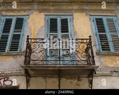 Flacher Blick auf einen julia-Balkon mit dekorativem schmiedeeisernem Geländer auf einem verwitterten Gebäude mit abgenutzten Holzläden; Athen, Griechenland Stockfoto