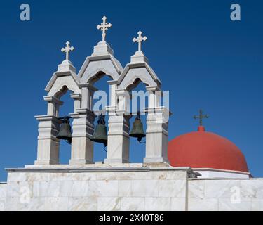 Architektonische Details von Glocken und Kreuzen und eine rote Kuppel auf dem Panagia Tourliani Kloster vor einem hellblauen Himmel in Mykonos, Griechenland Stockfoto