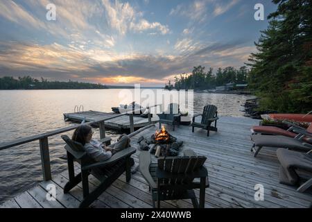 Frau sitzt in einem adirondack-Stuhl auf einer Seeseite vor einer Feuerstelle und beobachtet den Sonnenuntergang in der Ferne; Lake of the Woods, Ontario, Kanada Stockfoto