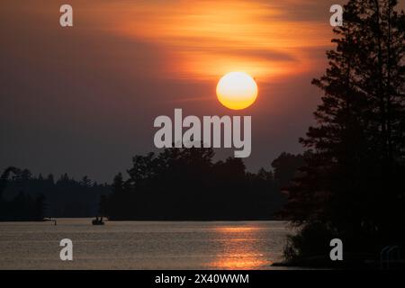 Große glühende Sonne untergeht am Himmel mit dunklen Wolken über einem See mit Silhouettenbäumen entlang der Küste und einem Sonnenstrahl, der auf dem Wasser reflektiert wird... Stockfoto