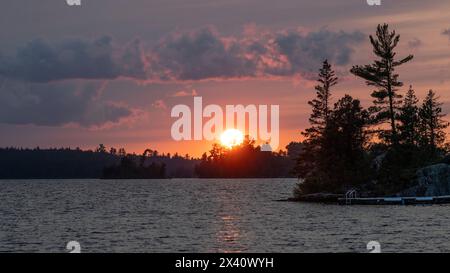 Dramatischer Blick auf die warme, glühende Sonne, die über dem Horizont mit einem See und Silhouetten an der Küste untergeht Stockfoto