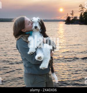 Mädchen zeigt Zuneigung zu ihrem Hund auf einem See bei Sonnenuntergang; Lake of the Woods, Ontario, Kanada Stockfoto