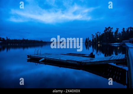 Blue Hour über einem ruhigen See mit einem Dock, der sich im Wasser spiegelt, und Bäumen entlang der Küste; Lake of the Woods, Ontario, Kanada Stockfoto