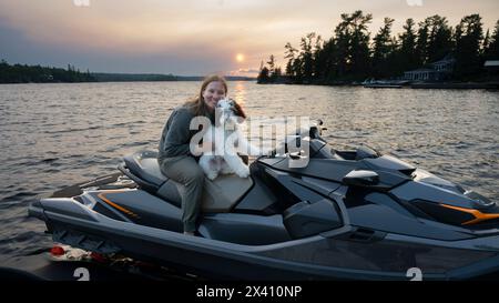 Eine junge Frau hält ihren Hund auf einem Wasserfahrzeug auf einem See bei Sonnenuntergang und posiert für die Kamera; Lake of the Woods, Ontario, Kanada Stockfoto