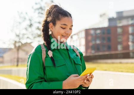 Fröhliches Teenager-Mädchen, das beim Gehen auf ihrem Smartphone schreibt Stockfoto