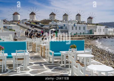 Touristen genießen die Uferpromenade von Mykonos, mit einer Restaurantterrasse im Vordergrund und den berühmten Windmühlen im Hintergrund; Mykonos, Griechenland Stockfoto