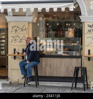 Mann sitzt auf einem Hocker unter einer Markise vor einem Café, das Gebäck verkauft; Athen, Griechenland Stockfoto