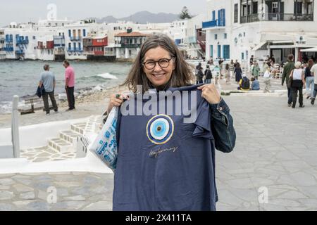 Weibliche Touristen halten ein Souvenir-T-Shirt in Mykonos, mit dem kleinen Venedig Einkaufsviertel entlang der Uferpromenade im Hintergrund Stockfoto