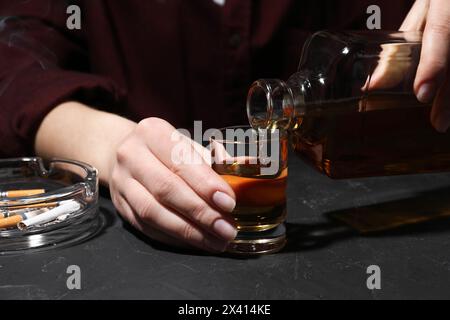 Alkoholsucht. Frau, die Whiskey aus der Flasche in Glas gießt, an einem dunklen, strukturierten Tisch, Nahaufnahme Stockfoto