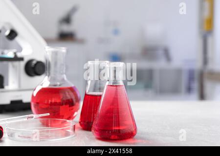 Laboranalyse. Verschiedene Kolben mit roter Flüssigkeit und Mikroskop auf hellgrauem Tisch im Innenbereich Stockfoto