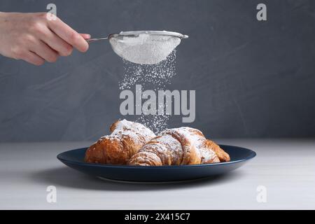 Frau mit einem Sieb, das Puderzucker auf Croissants streut, am weißen Holztisch, Nahaufnahme Stockfoto