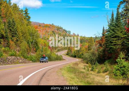 Ländlicher Highway mit Kurven und Hügeln und einem Fahrzeug und Herbstfarben an einem sonnigen Tag mit blauem Himmel im Norden von Minnesota. Stockfoto