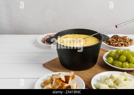 Fonduekanne mit leckerem geschmolzenem Käse, Gabel und verschiedenen Snacks auf weißem Holztisch, Platz für Text Stockfoto