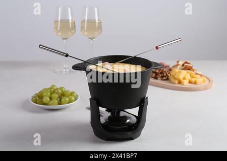 Fonduekanne mit leckerem geschmolzenem Käse, Gabeln, Wein und verschiedenen Snacks auf weißem Tisch Stockfoto