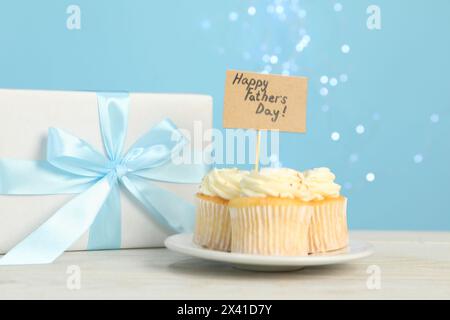 Alles Gute zum Vatertag. Leckere Cupcakes mit Grüßen und Geschenkbox auf hellem Holztisch, Nahaufnahme Stockfoto