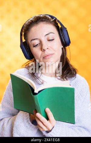 Eine fröhliche Frau, die sich über Bücher und Musik dreht, die Freude am Lesen vermittelt, Studiohintergrund. Geek liest Roman und hört Lieder im Kopfhörer, zeigt Wertschätzung für Literatur Stockfoto