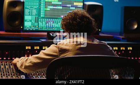 Fröhlicher, energischer Toningenieur tanzt im professionellen Studio auf seiner eigenen Musikkreation und bearbeitet eine Aufnahme auf einer Mischkonsole und daw-Software. Sounddesigner verarbeitet Titel. Kamera A.. Stockfoto