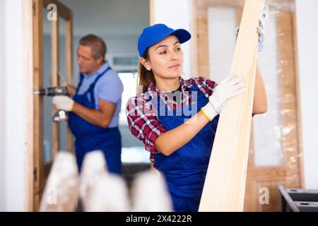 Positive lateinische Zimmerlerin in Uniform, die Holzbohlen im Innenbereich hält Stockfoto