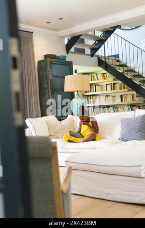 Afroamerikanerin in gelbem Kleid, liest zu Hause auf weißer Couch Stockfoto