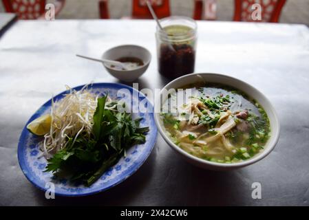 Vietnamesische Phở enthält eine Schüssel Reisnudeln in Brühe mit Rindfleisch und einen Teller mit Kräutern und Limette. Stockfoto