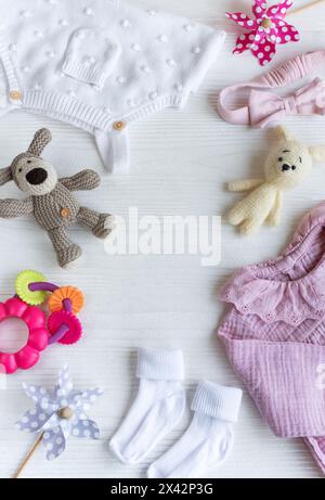 Set mit pinkfarbenen Kleidern und Accessoires für Neugeborene. Gestricktes Spielzeug Hase und Hund, Strampler, Socken und Handband auf weißem Holzhintergrund. Babydusche Stockfoto