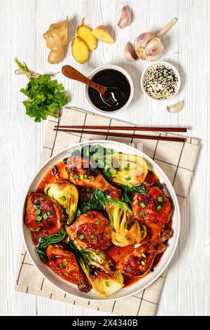 Gebratene asiatisch glasierte Hähnchenschenkel und Drumsticks mit gedämpftem Bok Choy in Schüssel auf weißem Holztisch mit Knoblauch, Ingwer, Sojasauce und Essstäbchen, Stockfoto