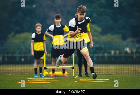 Teenage Boy beim Training mit dem jungen Fußballtrainer. Jugendfußballspieler springt auf dem Sporttrainingsfeld über eine Reihe von Hürden. Sportliche Stärke und Stockfoto