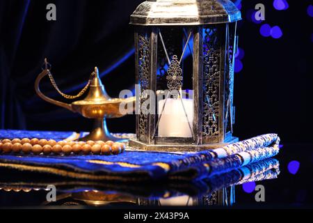 Arabische Laterne, misbaha, Aladdin Zauberlampe und gefaltete Gebetsmatte auf Spiegeloberfläche gegen verschwommenes Licht bei Nacht Stockfoto