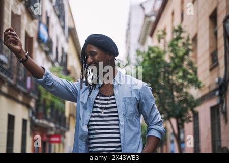 Afroamerikanischer Mann mit Dreadlocks und einer Baskenmütze, die die Hand hebt, fröhlich und mit geschlossenen Augen mitten in einer Stadtstraße. Stockfoto