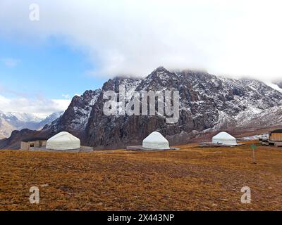 Jurten auf einem Bergplateau mit schneebedeckten Gipfeln im Hintergrund, unter bewölktem Himmel. Stockfoto