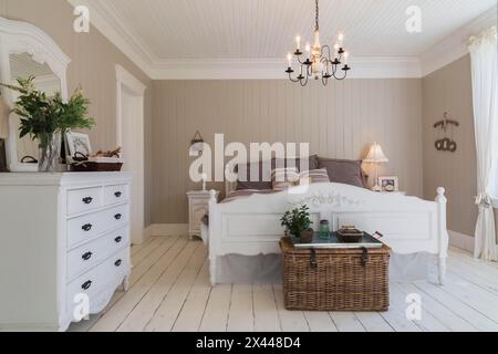 Kingsize-Bett mit hellbraun und beige gestreiften Bettüberwürfen, weiß lackiertem Kopfteil und Fußteil aus Holz mit geschnitzten Details, Kommode in Master Stockfoto