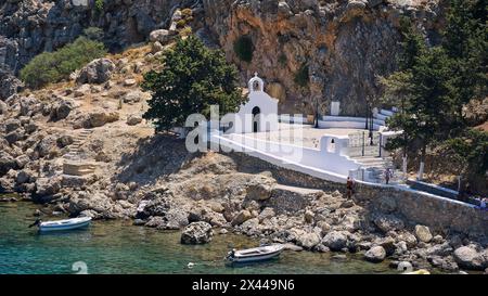 Eine weiße Kirche am Mittelmeer an einem felsigen Ufer neben geparkten Booten, St. Paul's Chapel, Paul's Bay, unterhalb der Akropolis von Lindos, Lindos Stockfoto