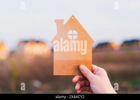 Die Hand des Mannes hält ein hölzernes Haus auf dem Hintergrund verschwommener Gebäude Stockfoto