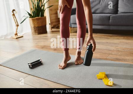 Eine abgeschnittene afroamerikanische Frau in Aktivkleidung, die auf einer Yogamatte steht und eine Flasche Wasser hält. Stockfoto