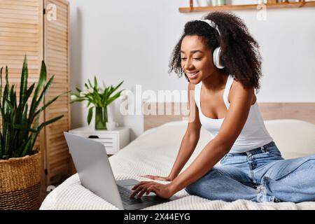 Lockige Afroamerikanerin in einem Tanktop, sitzt auf einem Bett und konzentriert sich intensiv darauf, einen Laptop-Computer in einem modernen Schlafzimmer zu benutzen. Stockfoto