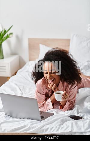 Eine lockige Afroamerikanerin im Pyjama liegt friedlich auf einem Bett und genießt morgens einen Laptop und eine Tasse Kaffee in ihrem Schlafzimmer. Stockfoto