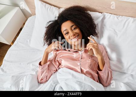 Lockige Afroamerikanerin im Pyjama, die auf dem Bett liegt und ein Papier mit nachdenklichem Ausdruck hält. Stockfoto