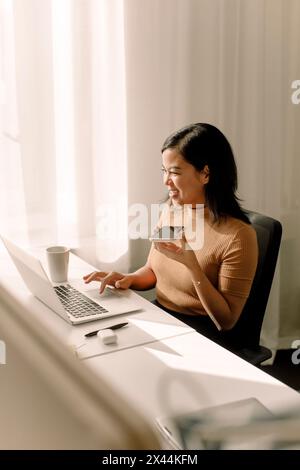 Glückliche Geschäftsfrau, die ihren Laptop benutzt, während sie über den Smartphone-Lautsprecher am Schreibtisch im Büro spricht Stockfoto