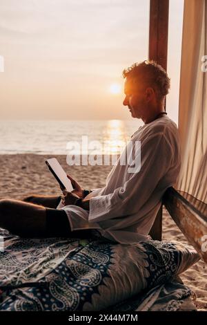 Seitenansicht eines Mannes, der einen Tablet-PC benutzt, während er im Urlaub am Strand in der Cabana sitzt Stockfoto