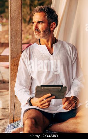 Ein reifer Mann, der einen Tablet-PC hält, während er im Urlaub am Strand in der Cabana sitzt Stockfoto