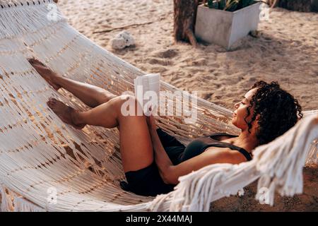 Eine Frau, die Buch liest, liegt in der Hängematte im Urlaub am Strand Stockfoto