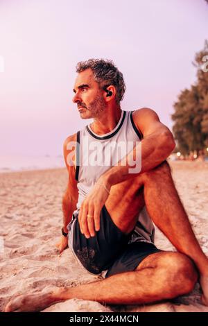 Reifer Mann, der sich dehnt, während er am Strand auf Sand sitzt Stockfoto