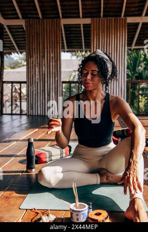 Frau hält Räucherstäbchen, während sie im Wellnessresort auf Yogamatte sitzt Stockfoto