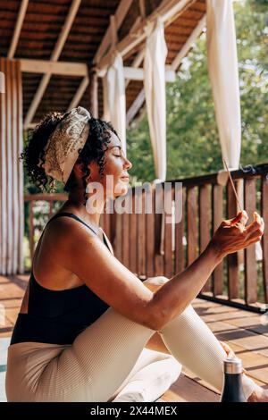 Seitenansicht einer Frau, die Meditation übt und Räucherstäbchen hält, während sie im Wellnessresort sitzt Stockfoto