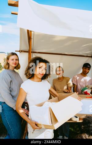 Porträt einer jungen Unternehmerin mit Pappkarton, während sie mit Kollegen in der Nähe des Zeltes im Gemeindezentrum steht Stockfoto