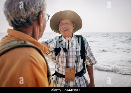 Glücklicher, schwuler Senior-Mann, der mit seinem Freund spricht, während er im Urlaub in der Nähe des Meeres am Strand stand Stockfoto