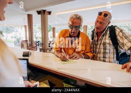 Ein schwules Senior-Paar füllt das Formular an der Rezeption im Hotel aus Stockfoto