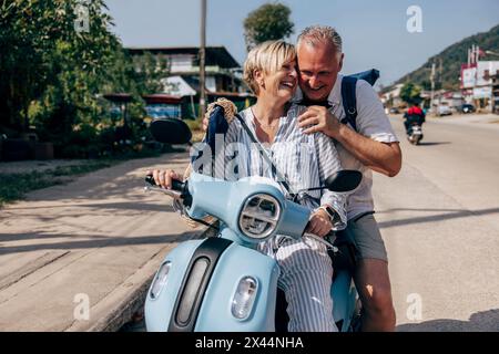Glückliche Seniorin, die auf dem Roller sitzt, mit einem Mann am Straßenrand Stockfoto
